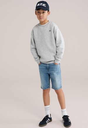 Hanorac gri, pantaloni scurți din denim albastru, pantofi sport negri cu dungi albe, șosete atletice albe și o șapcă închisă la culoare cu inscripția „N.Y.C.”.