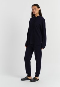 Felpa blu marinara a maglia con tasca frontale, abbinata a pantaloni blu marinara a gamba tapered e sandali neri con dettaglio in oro sulla fibbia.
