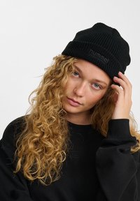 Beanie nera a maglia con texture a coste, con un logo sulla parte anteriore. Indossata da una persona con capelli lunghi e ricci, su uno sfondo semplice.