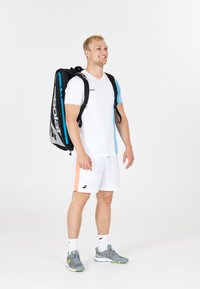 Giovane uomo che indossa abbigliamento sportivo bianco e sneakers grigie, con una borsa da tennis nera e blu sulla schiena, sorridente e in piedi.