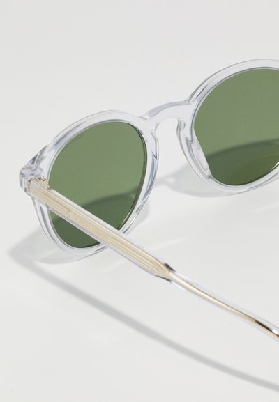 Ochelari de soare rotunzi, transparenți, cu lentile verzi. Au un cadru transparent și accente metalice aurii pe brațele templelor, cu un design texturat.