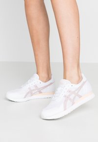 Vita atletiska sneakers i mesh och gummi, med rosa accenter och tre ränder på sidorna. Minimalistisk design med snörning.