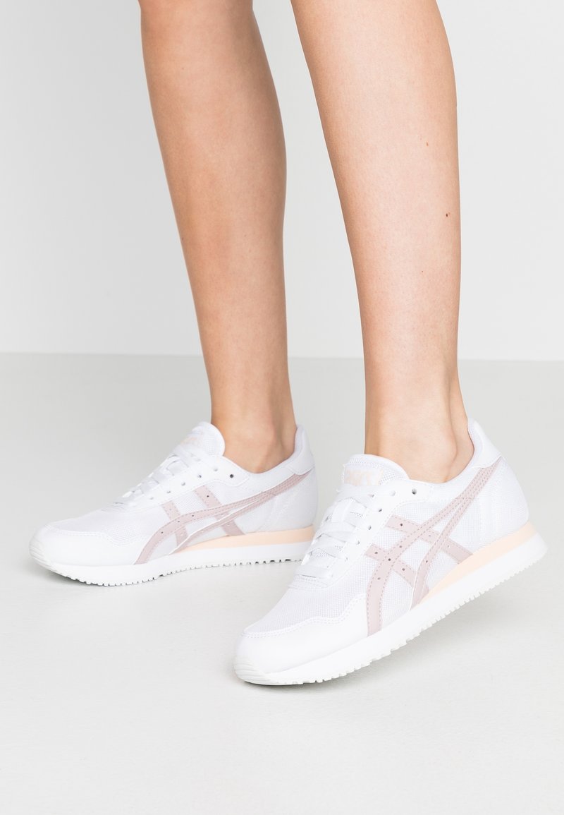 Vita atletiska sneakers i mesh och gummi, med rosa accenter och tre ränder på sidorna. Minimalistisk design med snörning.