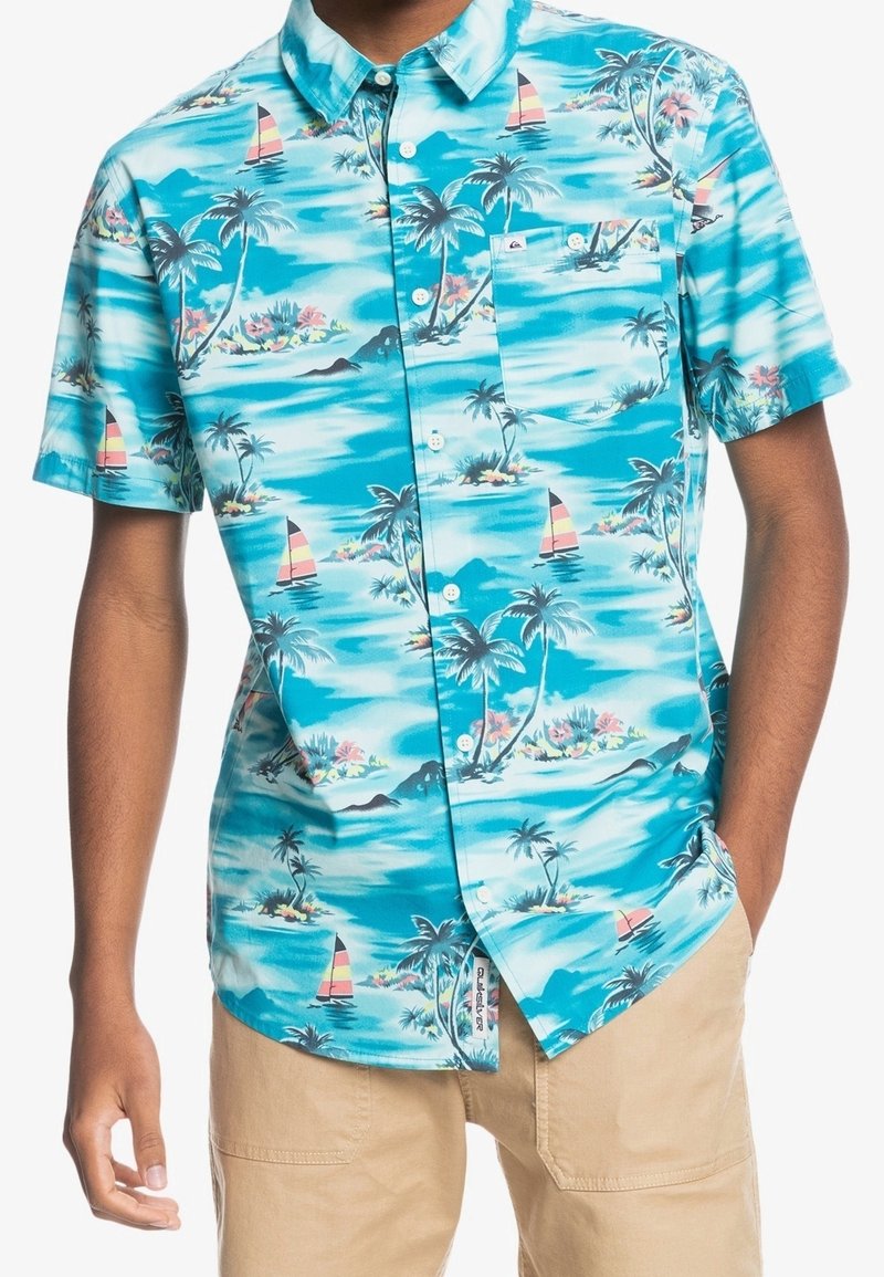 Camisa de manga corta con botones en azul claro con patrón tropical de palmeras y veleros. Cuenta con un bolsillo y un corte relajado. Material de algodón.