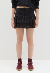 Minigonna nera con paillettes, caratterizzata da un design a strati con volant, vita aderente e tessuto trasparente, abbinata a scarpe da ginnastica bordeaux e calzini.