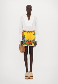 White, textured blouse with a deep V-neck, paired with a yellow mini skirt featuring colorful floral patterns and a drawstring. Holding a brown handbag.