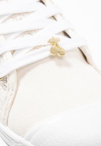 Baskets en toile beige clair avec des lacets blancs, ornées d'un petit accent en or en forme de cerise près des lacets et d'une semelle en caoutchouc texturée.