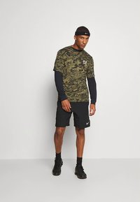 Kurzarm-T-Shirt mit Tarnmuster in Grün und schwarzen langen Ärmeln, kombiniert mit schwarzen Shorts und schwarzen Sportschuhen. Sichtbare Logos von Under Armour und Nike.