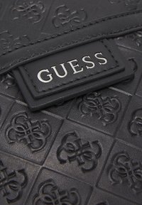 Černá embosovaná kožená kabelka s texturovaným vzorem. Obsahuje výraznou stříbrnou jmenovku "GUESS" na hladkém obdélníkovém poli.