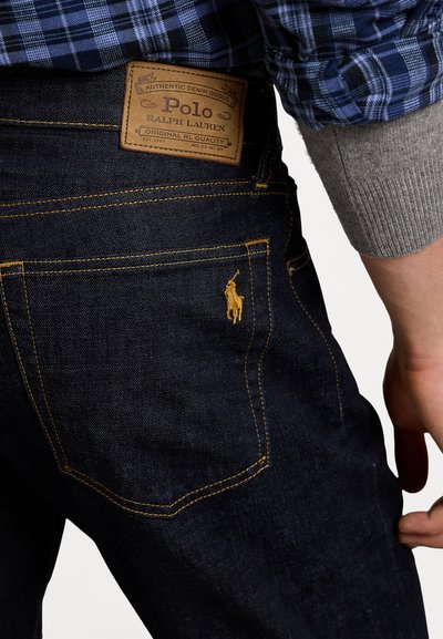 Jeans in denim blu scuro con una toppa in pelle beige e cuciture gialle. Emblema di un cavallo ricamato sulla tasca posteriore. Design classico.