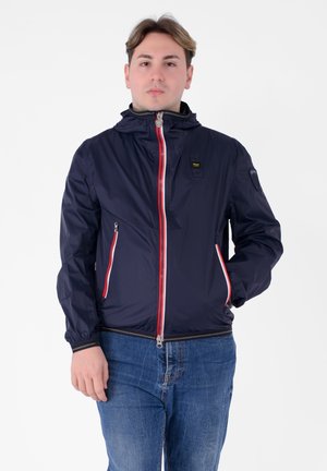 Navyblauwe lichtgewicht jas met een rode rits en accenten, voorzien van een capuchon, elastische manchetten en zijzakken. Soepele textuur met een strakke uitstraling.