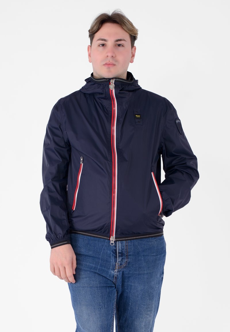 Giacca leggera blu navy con cerniera rossa e dettagli in rosso, dotata di cappuccio, polsini elasticizzati e tasche laterali. Texture liscia con un design elegante.