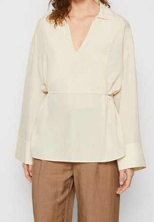 Helle Cremebluse mit V-Ausschnitt, langen Ärmeln und taillierter Passform. Hergestellt aus glattem Stoff, mit minimaler Textur und klaren Linien.