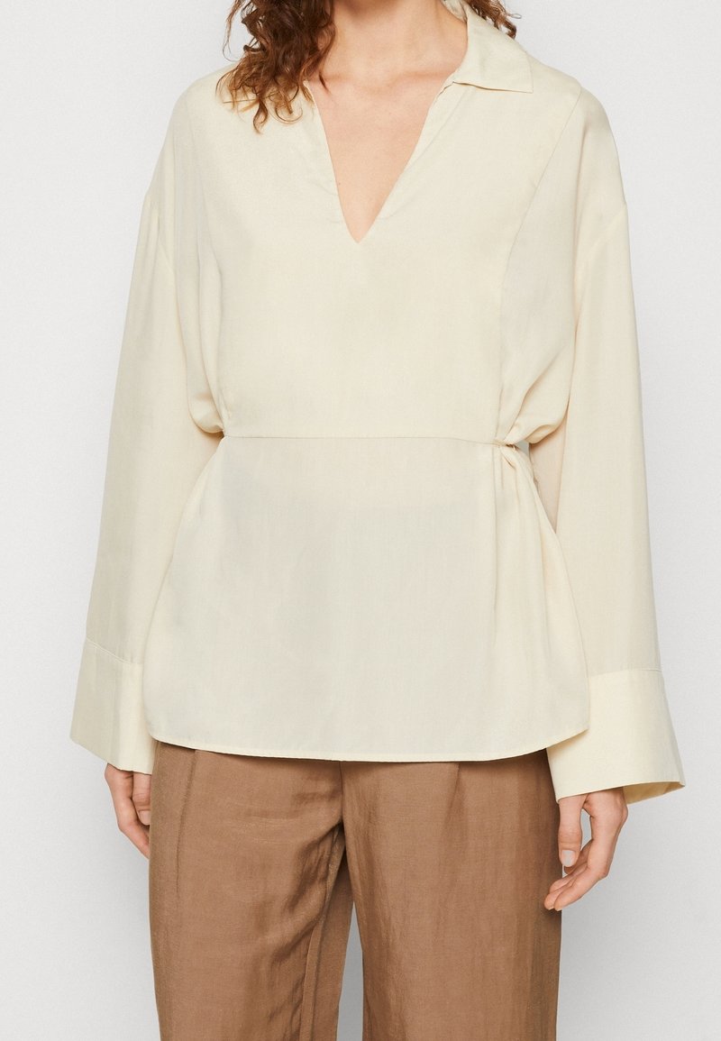Blouse crème clair avec un décolleté en V, des manches longues et une taille cintrée. Fabriquée en tissu lisse, présentant une texture minimale et des lignes épurées.