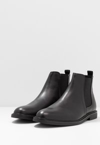 Botines Chelsea de cuero negro con paneles laterales elásticos, punta redondeada y suela de goma plana. Textura suave y detalles de diseño minimalistas.