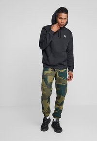 Felpa nera con cappuccio, tasca frontale e logo bianco, abbinata a pantaloni mimetici verdi e marroni. Scarpe da ginnastica nere completano l'outfit.