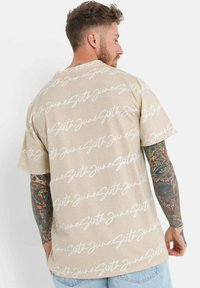 Beige kortermet t-skjorte med hvit kursiv tekstmønster. Myk stoff, løs passform med rund hals, og synlige tatoveringer på armen.