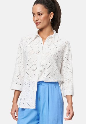 Chemise blanche brodée avec un col boutonné, ornée de motifs en œillet complexes et de manches trois-quarts, portée avec un pantalon bleu clair.