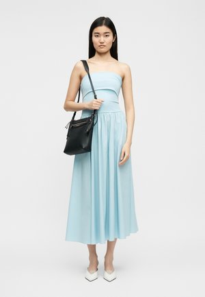 Femme portant une robe midi bleu clair sans bretelles, des chaussures blanches pointues, et un sac à bandoulière noir, debout devant un fond uni.