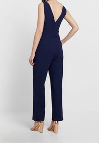 Marinblå bodysuit med V-rygdesign, ärmlös snitt och rak passform. Materialet ser slätt ut med en subtil glans. Bärs med högklackade skor.