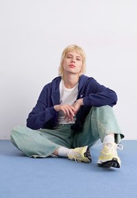 Sudadera de marina con logo blanco, camiseta blanca, jeans holgados verde claro, zapatillas amarillas y negras, sentado sobre una superficie azul, fondo neutro.