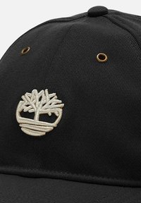 Cappello nero con un logo ad albero ricamato beige sul davanti. Tessuto liscio con una finitura textured; include occhielli in metallo dorato.