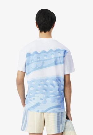 Homme aux cheveux noirs courts, debout de dos, portant un T-shirt blanc et bleu à motifs et un short rayé, tenant un sac assorti.