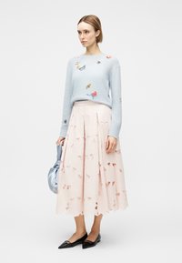 Γυναίκα που φοράει ανοιχτό μπλε πουλόβερ με μικρά κεντητά έντομα και πεταλούδες, ανοιχτό ροζ μίντι φούστα με κοψίματα, μαύρες μπαλαρίνες, κρατώντας ένα μπλε τσάντα.