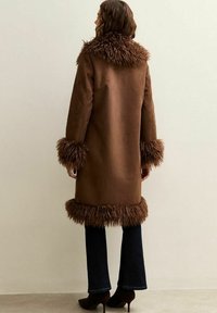 Bruine jas met een pluizige, harige bontkraag en manchetten. De jas heeft een gladde oppervlakte, is knie-lange en is gecombineerd met uitlopende jeans.