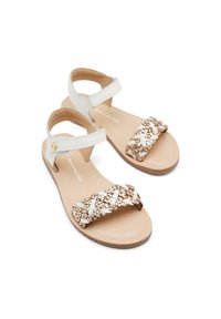Sandalias con suela beige, tira de tobillo ajustable blanca y una banda trenzada metálica dorada y blanca en la parte delantera. Diseño plano, acabado texturizado.