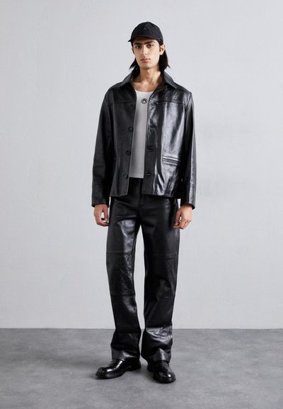 Μαύρο δερμάτινο μπουφάν με κούμπωμα και γκρι πουκάμισο από κάτω, συνδυασμένο με μαύρο δερμάτινο παντελόνι με υφασμάτινες λεπτομέρειες και μαύρα παπούτσια.