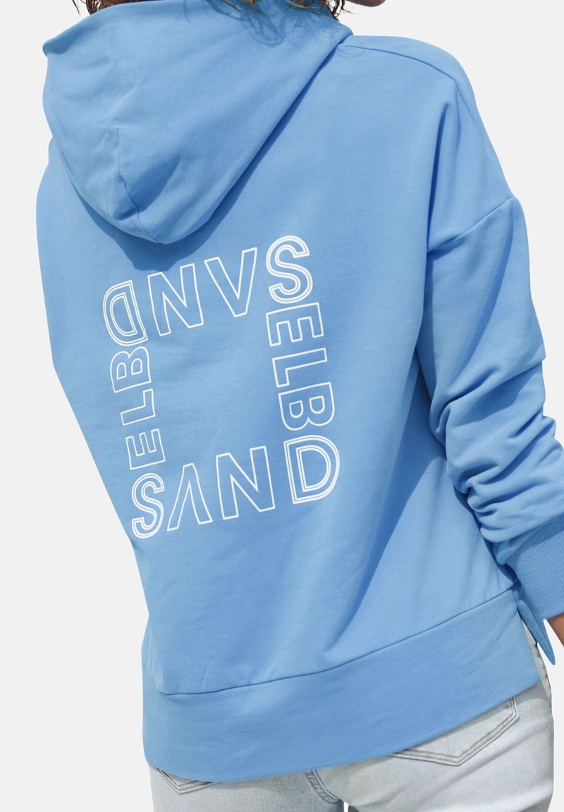 Personne portant un sweat à capuche bleu clair avec le mot « SELBD SAND » imprimé en lettres blanches au dos en forme carrée.