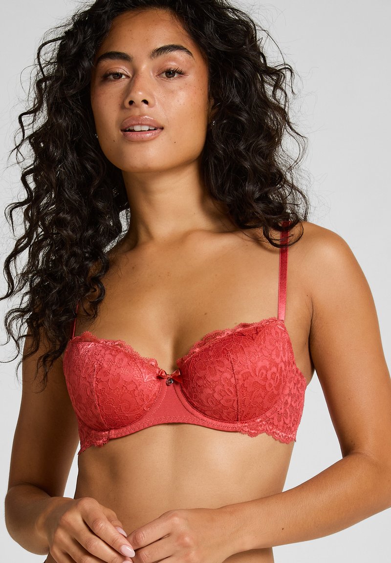 Frau mit langen lockigen Haaren, die einen roten Spitzen-Balconette-BH trägt und vor einem schlichten hellgrauen Hintergrund posiert.