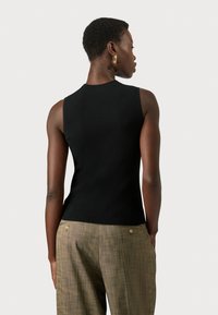 Zwarte geribbelde mouwloze top met een aansluitend ontwerp, gezien vanuit de rug, gecombineerd met lichtbruine geruite broek met gouden knopen.