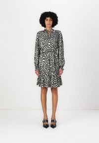 Robe à motifs noir et blanc avec des manches longues, un col et une taille élastique. L'ourlet présente un détail volants. Portée avec des escarpins noirs.