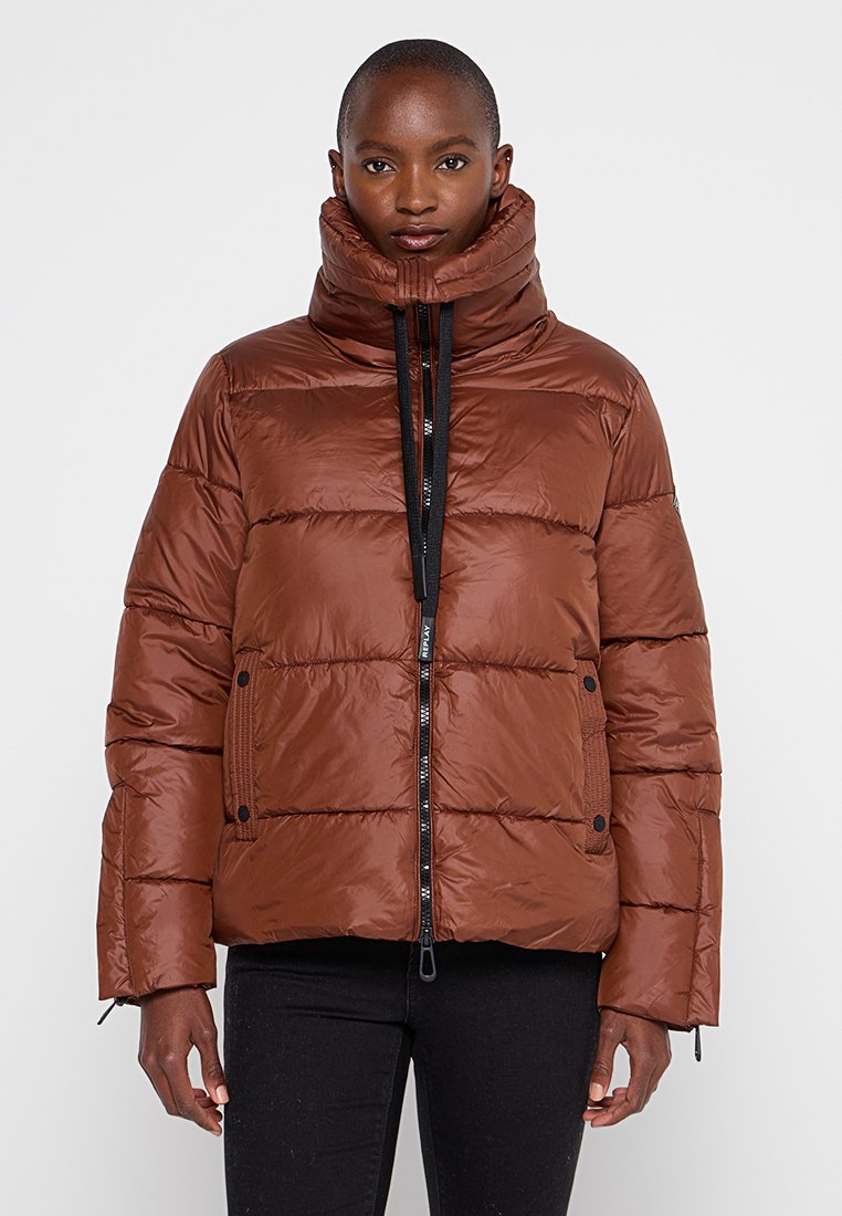 Persona che indossa un giubbotto trapuntato marrone con colletto alto e chiuso, in piedi contro uno sfondo chiaro e uniforme.