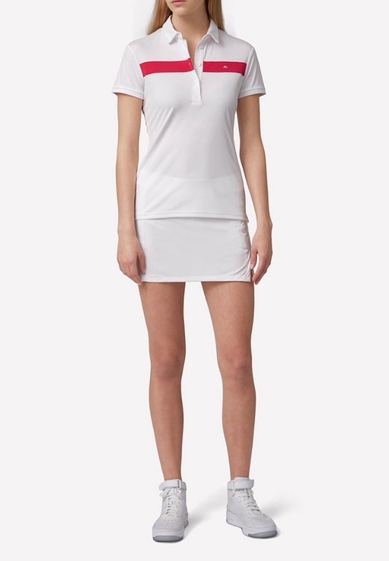 Biała koszulka polo z różowym poziomym paskiem, zestawiona z dopasowaną białą spódnicą. Materiał wydaje się gładki i lekki.