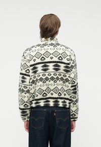 Pūkinio audinio striukė su juoda ir balta geometrine rašte, apvaliu apykakle ir laisvu siluetu. Dėvima su tamsiai mėlynomis džinsinėmis kelnių.