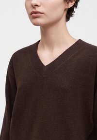Jeune personne portant un pull en maille marron foncé à col en V, montrant le cou, la mâchoire et le haut du torse sur un fond clair uni.