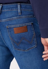 Jeans de mezclilla azul con un parche de cuero marrón en el bolsillo trasero, con costuras en contraste y un patrón de zigzag distintivo.