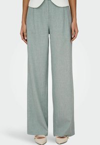 Femme portant un pantalon large gris clair et des ballerines crème pointues, debout droite avec les bras détendus le long du corps.