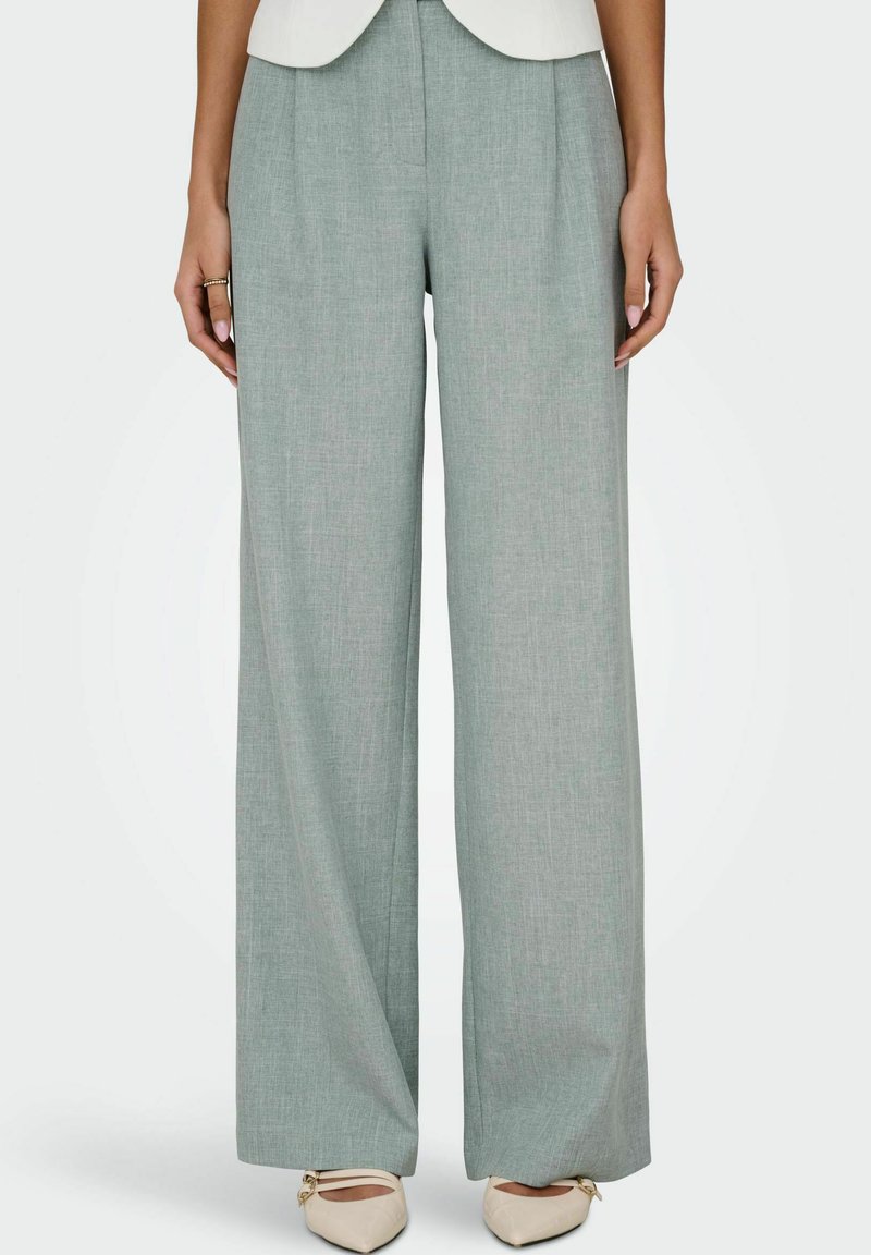 Femme portant un pantalon large gris clair et des ballerines crème pointues, debout droite avec les bras détendus le long du corps.