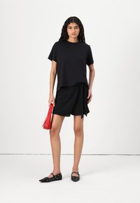 Sort T-shirt med korte ærmer, lagdelt design. Sort indpakningsnederdel, rød skuldertaske og sorte flade sko med cirkulære udskæringer. Minimalistisk stil.