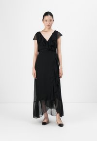 Frau steht in einem schwarzen, gerafften Wickelkleid mit durchsichtigen Ärmelchen und einem asymmetrischen Saum, trägt zweifarbige Ballerinas.
