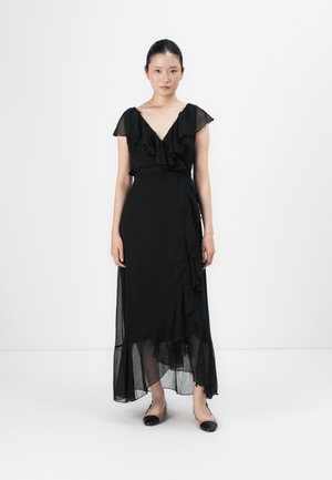 Kvinne står i en svart volang-innsvøpt kjole med gjennomsiktige cap sleeves og en asymmetrisk søm, iført bicolore flate sko.