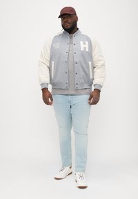 Jachetă bomber gri și crem, cu nasturi tip snap, având un logo "H", purtată peste un tricou gri, blugi albaștri deschis și adidași albi.