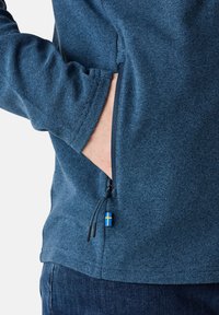 Blått texturerat tygjacka med en dragkedjeficka och en dragflik, prydd med en liten svensk flagga.