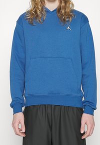 Osoba v modré mikině Jordan s přední kapsou a černých kalhotách, ukazující pouze trup a ruce na bílém pozadí.