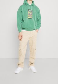 Groene hoodie met een grafische print, gecombineerd met beige cargobroek en witte sneakers. De hoodie heeft een trekkoord en een zachte textuur.