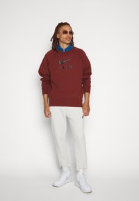 Kastanjefärgad Nike-hoodie med svart logotyp, ihop med vita joggers och vita sneakers. Modellen bär solglasögon och en blå skjorta under.